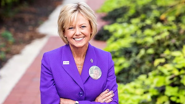A video screen shot of stroke survivor, Ann, a white woman smiling and standing with arms crossed in an outdoor setting