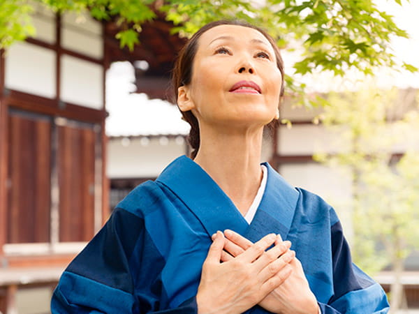 Una mujer japonesa de edad avanzada, vestida con un kimono azul, tiene las manos apoyadas sobre el pecho con un gesto de sanación mientras mira hacia el cielo.