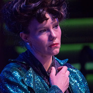 Maggie Whittum is wearing a teal sequined jacket and has an expression of sadness on her face while acting out a scene in a play. Maggie Whittum is wearing a teal sequined jacket and has an expression of sadness on her face while acting out a scene in a play.