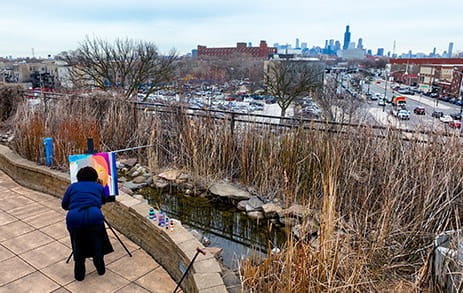 Stroke survivor, Minnie, is painting on a canvas in a park overlooking city buildings and streets.