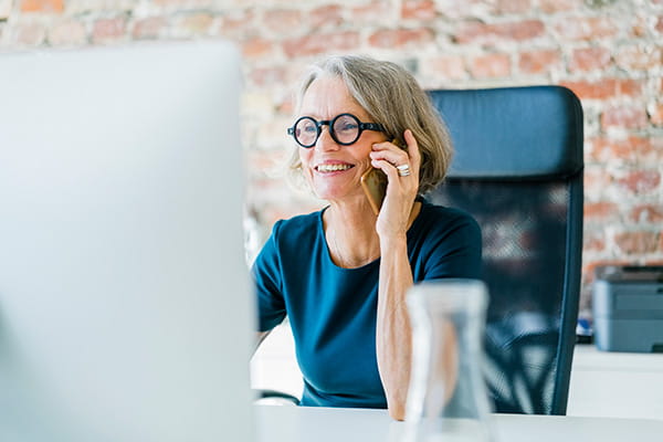 A senior woman is sitting at her desk and talking on a mobile phone.