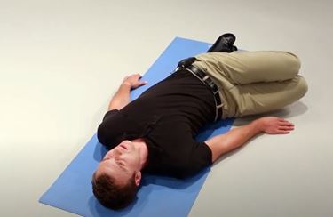 A White man is lying flat on his back on a mat with his knees bent and rotated to the right.