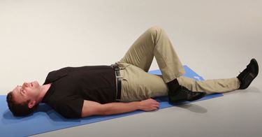 A White man is lying flat on his back on a mat with his left leg bent and crossed over his outstretched right leg.