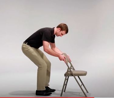 A White man is leaning over in a partial squat with this hands resting on the back of a folding chair for support.