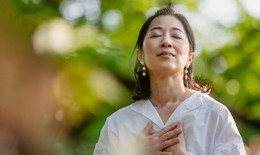 A woman is closing her eyes, doing a breathing exercise and meditating in nature.