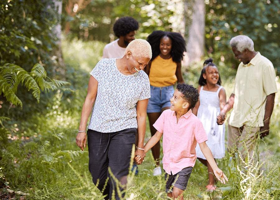A senior Black woman and man are walking with their multigenerational family along a nature trail through trees.